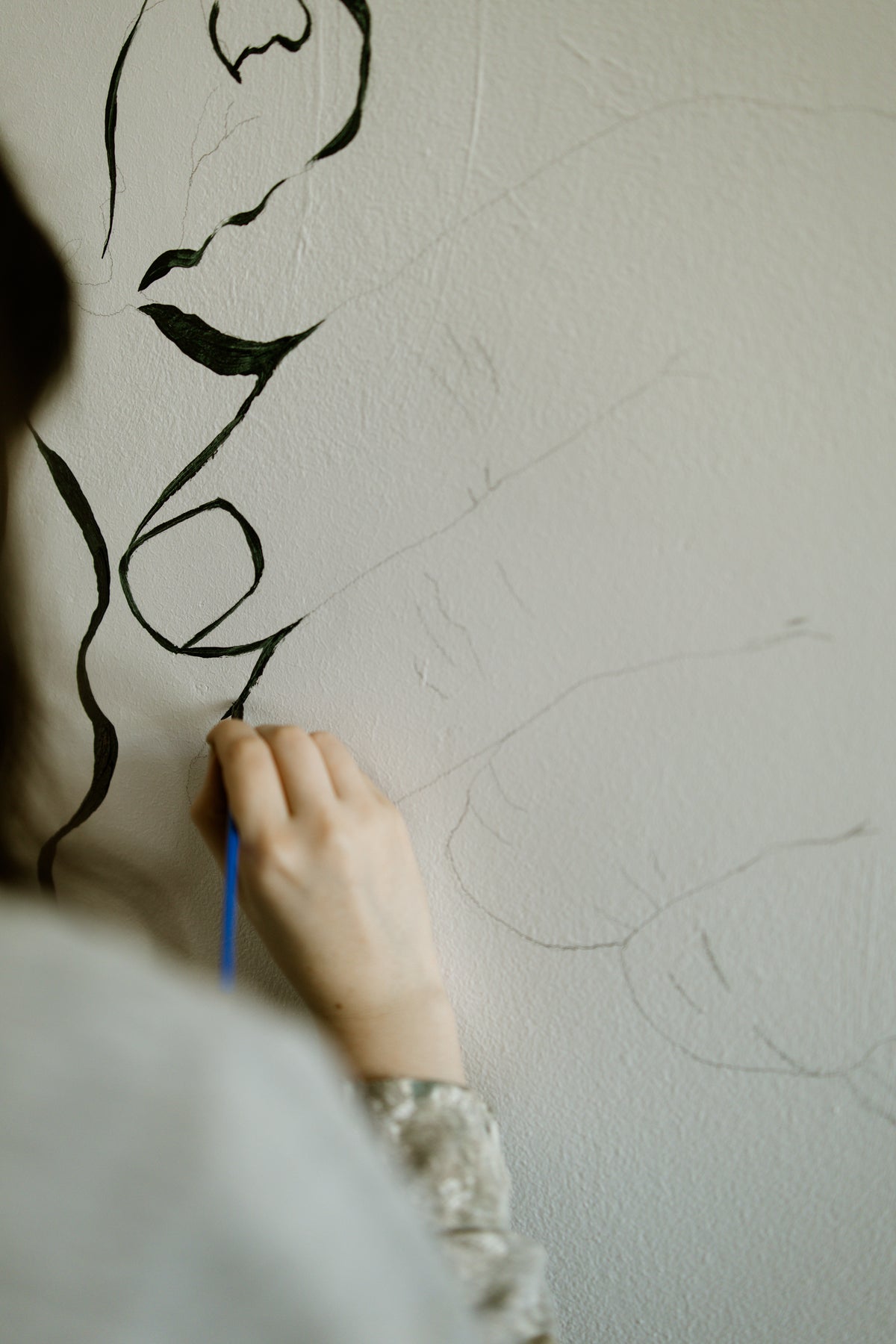 A close-up of a person's hand holding a blue paintbrush and applying black paint to a textured plaster wall, creating an abstract line drawing.